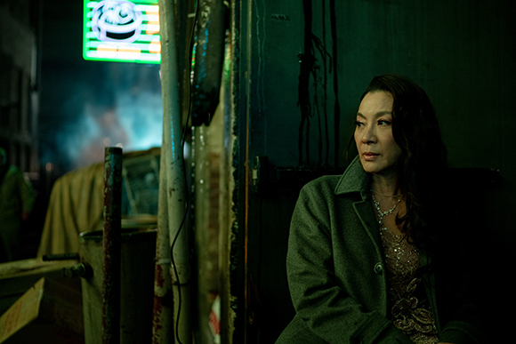 A woman in a grey coat looks pensively into the distance in a dark, narrow alleyway.