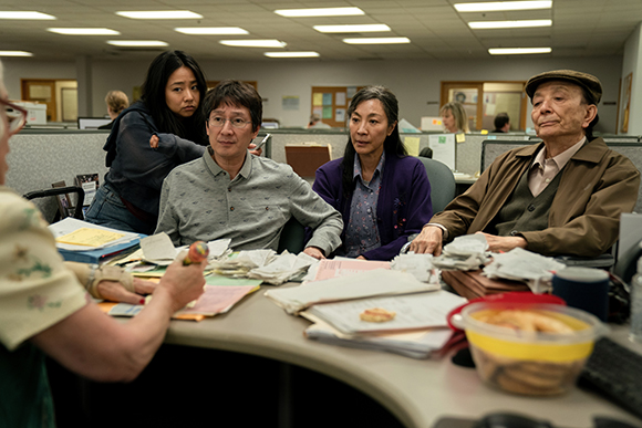 Four people sit around a desk cluttered with papers and documents in an office.