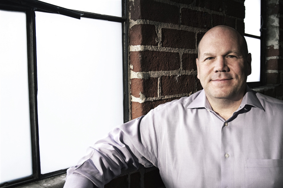 A man in a light purple button-down shirt sits in front of a brick wall and a large window.