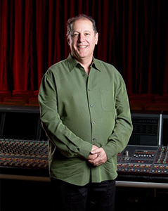 A smiling man in a long-sleeved green button-down shirt stands in front of a mixing console and red curtains.