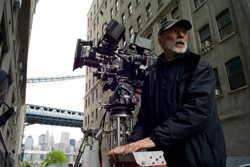 A man in a black jacket stands beside a large professional film camera on a tripod in a city.