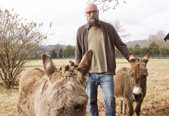 A bearded man in a cardigan and jeans stands between two donkeys in a grassy field.