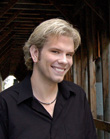 A smiling man with blond hair wearing a black button-down shirt and a thin necklace.