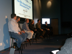 Six people sit in a row on stools facing a large projection screen in a dark room.