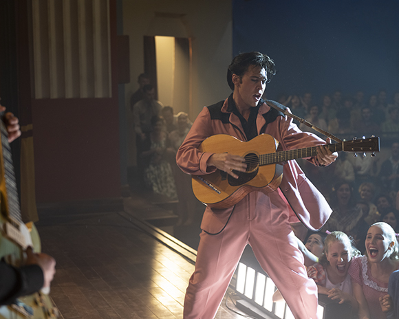 A man in a pink suit plays an acoustic guitar on a stage in front of a crowd.