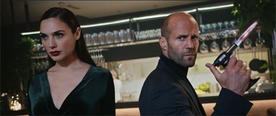 A woman in a dark green velvet dress and a man in a black turtleneck stand back-to-back, holding a knife.