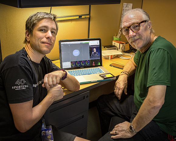 Two men sit at a desk with an open laptop displaying video editing software.