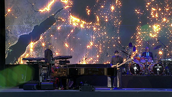 A band performs on stage with a large digital backdrop of glowing city lights at night.