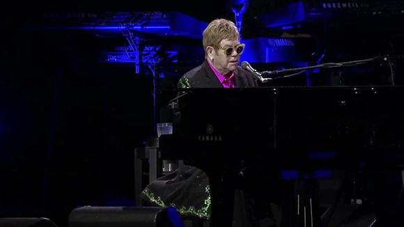 A man with blonde hair and yellow sunglasses plays a black Yamaha grand piano on a dark stage.