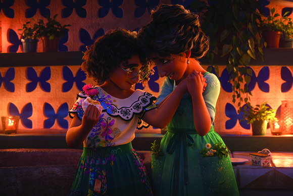 Two young girls in colorful dresses smile and hold hands in a warm, candlelit courtyard.