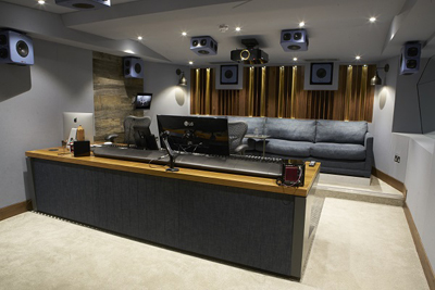A modern recording studio featuring a mixing desk, computer monitors, and a blue sofa with surround sound speakers.