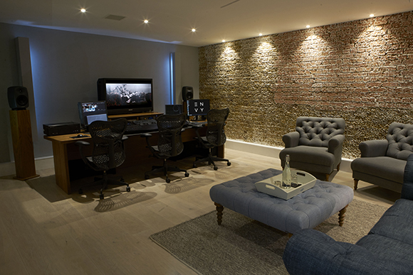 A media studio with a large monitor, mixing desk, and office chairs facing a brick wall, with a lounge area in front.