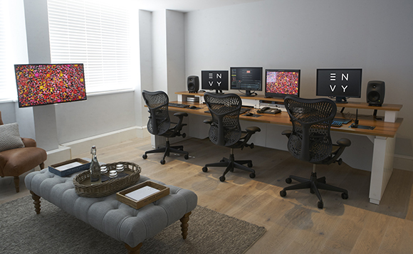 Three workstations with black mesh chairs and monitors sit along a long wooden desk in a bright room.