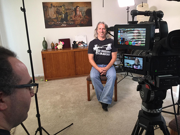 A man in a Dungeons & Dragons t-shirt sits on a stool being filmed by a professional video camera setup.