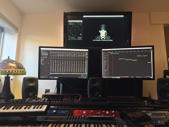 Music studio with three computer monitors, studio monitors, a MIDI keyboard, and audio interfaces.