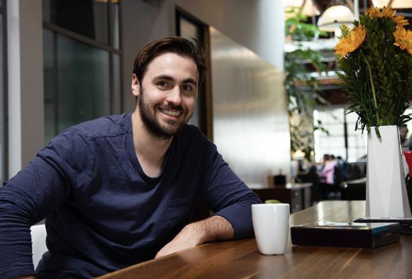 A smiling man with dark hair and a beard wearing a navy long-sleeve shirt, sitting at a wooden table.