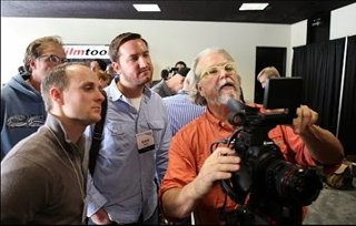 A man in an orange shirt operates a professional video camera while several men watch him.