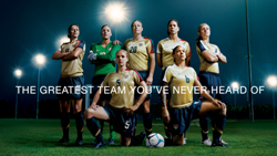 Seven female soccer players in gold and blue uniforms pose on a grass field under bright stadium lights.