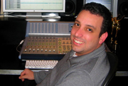 A man in a grey sweater sits in front of a large audio mixing console and computer monitors.