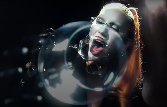 A person with red hair and glasses blowing a large, translucent bubble through a glass tube.
