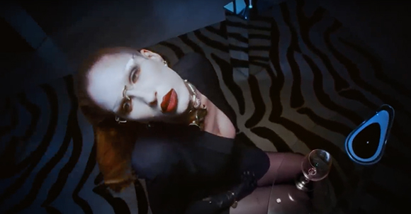 A person with heavy eye makeup and red lipstick reclines against a zebra-patterned surface.