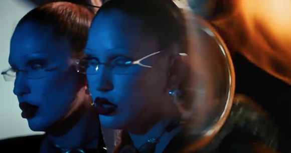 A woman with dark lipstick and glasses reflected in a mirror under blue light.