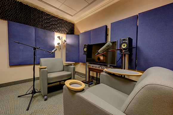 A recording studio with two grey armchairs, microphones, monitors, and blue acoustic wall panels.