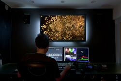 Person sitting in a dark room viewing a large screen with golden particles and two smaller computer monitors.