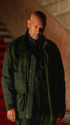 A bald man wearing a dark green utility jacket looking down with a serious expression.