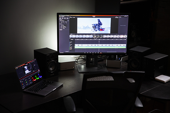 A large computer monitor displaying video editing software flanked by studio monitors and a laptop on a desk.