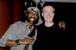 A man in a patterned shirt and hat holds a guitar while standing next to a man in a black shirt.