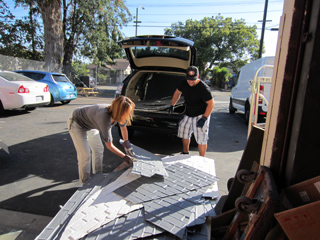 A person unloads stacks of grey roof shingles from the open trunk of a black car.