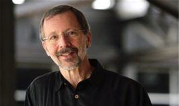A man with short brown hair, glasses, and a graying beard wearing a black shirt, smiling against a blurred background.