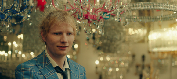 A man with blond hair wearing a blue plaid blazer in front of sparkling crystal chandeliers.