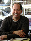 A smiling man in a dark long-sleeved shirt sits at a desk with photos pinned to the wall behind him.