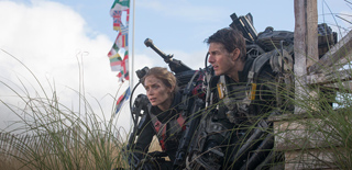 Two people in futuristic combat gear crouch behind tall grass under a cloudy sky.