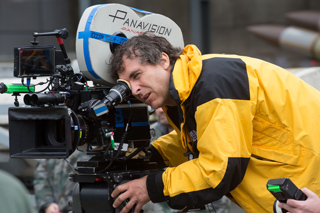 A man in a yellow and black jacket looks through the viewfinder of a large Panavision film camera.