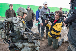 A camera operator films an actor wearing heavy, weathered mechanical armor on a film set.