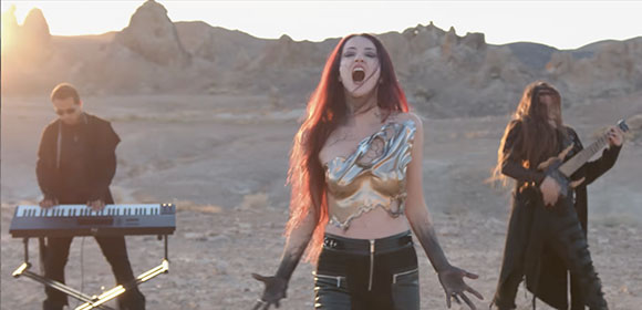 A woman sings passionately in a desert while a man plays a keyboard and another plays guitar.