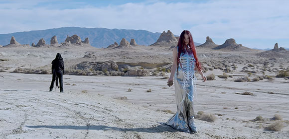 A person in a long silver dress walks through a desert, while another figure stands in the distance.