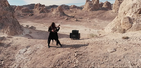 A person in black clothing plays a guitar next to an amplifier in a rocky desert landscape.