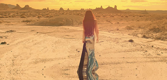 A person in a long, flowing dress walks away through a vast, sandy desert toward distant rock formations.