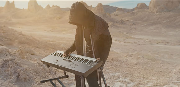 A person wearing a dark hooded cloak plays a musical keyboard in a rocky, sunlit desert landscape.
