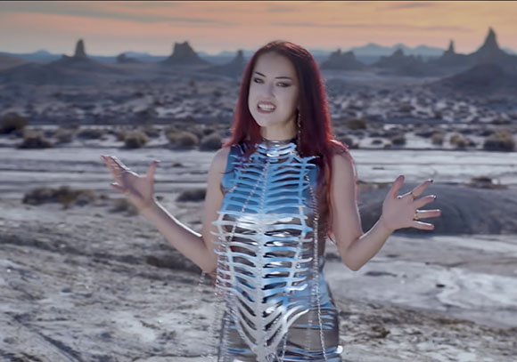 A woman with long red hair wears a silver metallic skeletal bodice in a desert landscape at sunset.