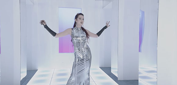 A woman in a long silver dress and black arm sleeves poses with arms outstretched in a white studio.
