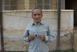 A man in a light grey shirt holds up a small black and white portrait of a person.
