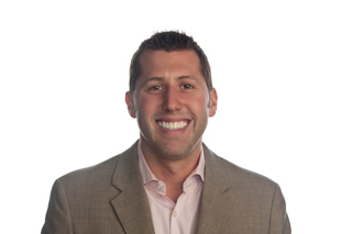 A man with short brown hair, wearing a light pink collared shirt and a tan suit jacket, smiles broadly against a white backgr