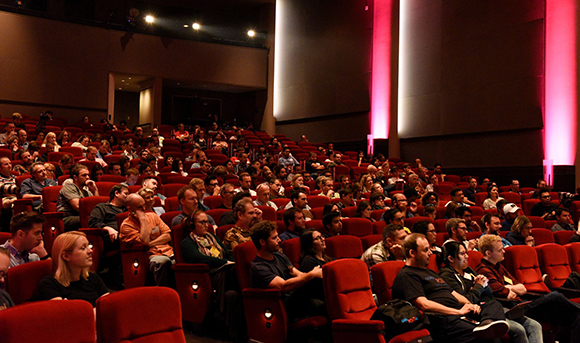 A large audience sits in rows of red velvet seats inside a darkened movie theater.