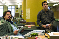 A man with glasses and a mustache sits at a desk while another man in a plaid shirt stands nearby in an office.
