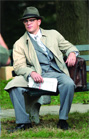 A man wearing a fedora, glasses, a grey suit, and a tan trench coat sits on a park bench.